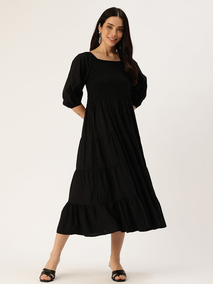 Woman in a black dress standing in a bright living room with plants and furniture. Black Solid Cuffed Sleeves Dress, midi dress, dress, women’s western wear, party wear dress, casual wear dress