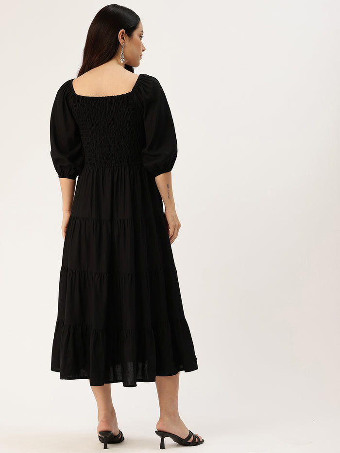 Woman in a black dress standing in a bright living room with plants and furniture. Black Solid Cuffed Sleeves Dress, midi dress, dress, women’s western wear, party wear dress, casual wear dress