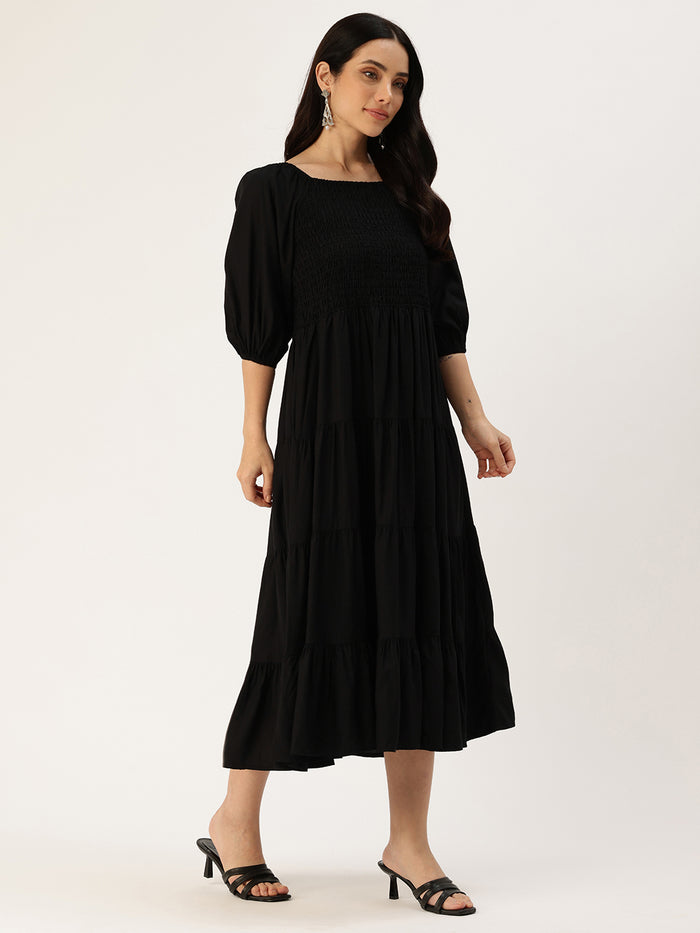 Woman in a black dress standing in a bright living room with plants and furniture. Black Solid Cuffed Sleeves Dress, midi dress, dress, women’s western wear, party wear dress, casual wear dress