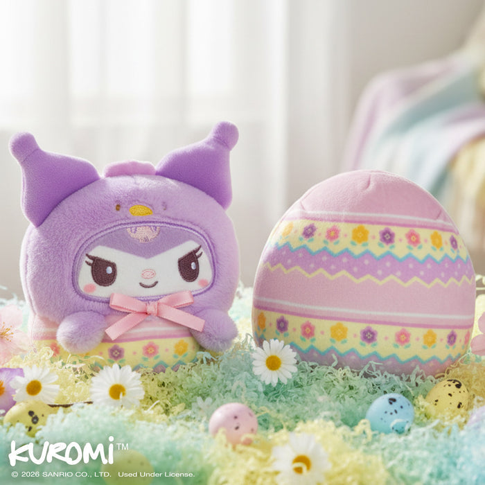 Easter Egg Kuromi 4" Reversible Plushie sitting on a table of pastel paper grass, flowers, and small chocolate eggs with a white curtained window and pastel blanket on a chair in the background