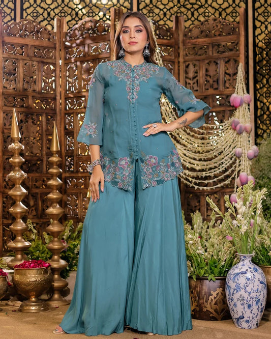 Woman in a teal traditional outfit with floral embroidery standing in a decorative setting.